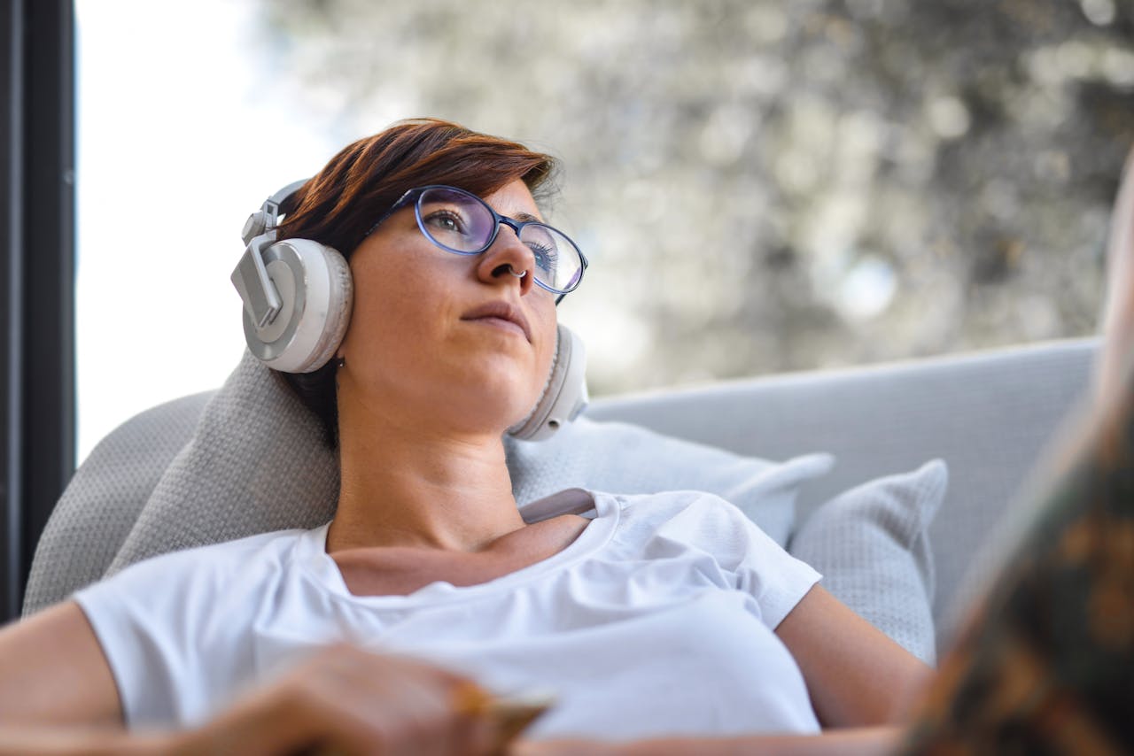 Woman in glasses listening to music with headphones, relaxing on a couch by a window.
