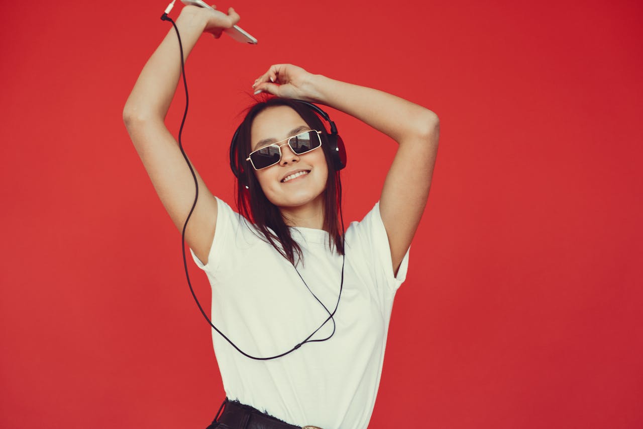 Optimistic positive millennial female in white top and stylish sunglasses listening to favorite playlist and looking at camera while dancing with raised arms in studio on red background