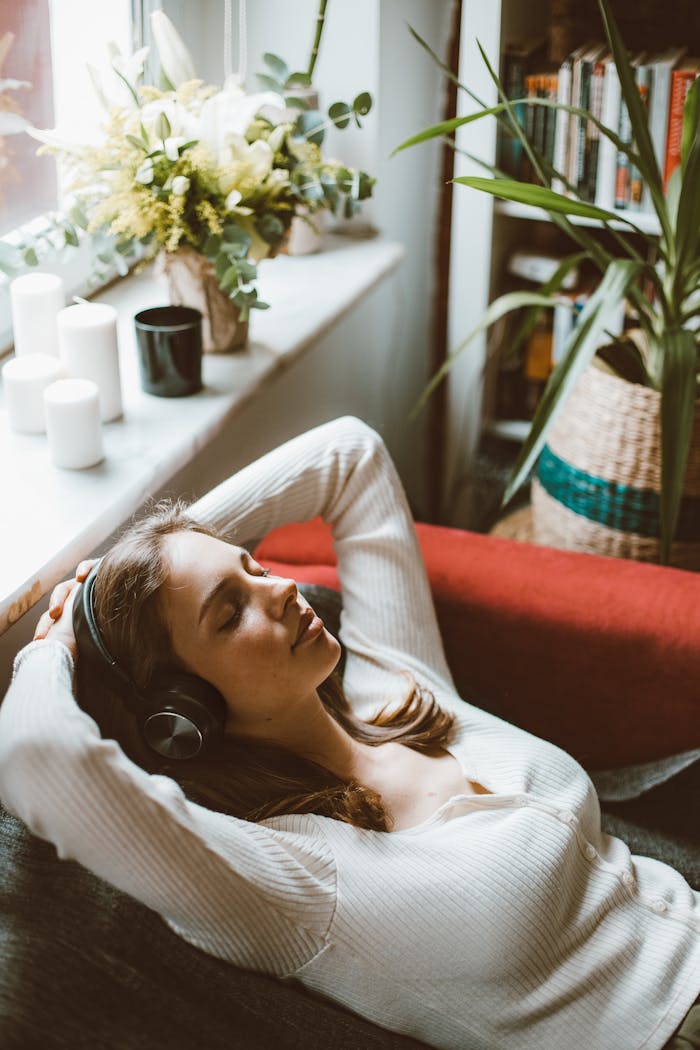 A woman in a white sweater relaxes in an armchair with headphones, enjoying music amidst a serene indoor setting.