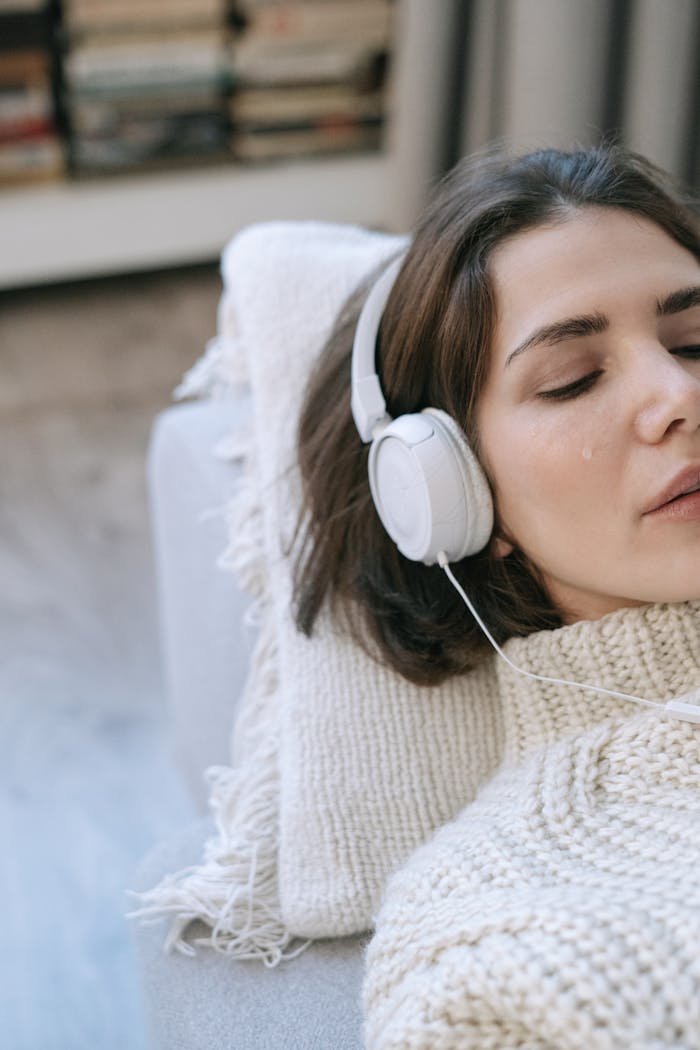 Young woman lying down indoors, wearing headphones and a knitted sweater, with tears on her face.